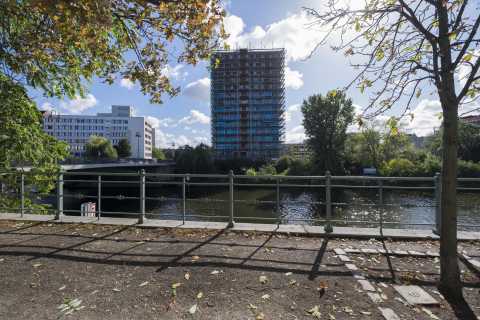 Fluss mit Uferweg, Baustelle am Hochhaus, Brücke und Bäume bei Sonnenschein.