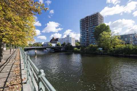 Fluss mit Uferweg, Brücke und hohen Gebäuden unter blauem Himmel.