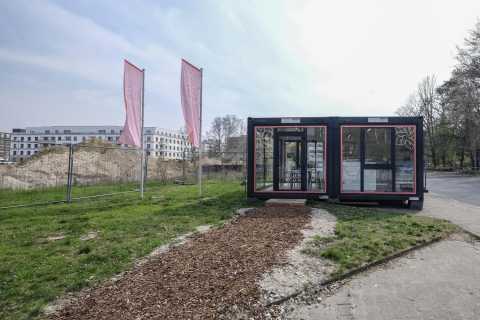 Zwei Container mit Glasfronten, davor Holzweg, daneben Baustelle und Fahnen.