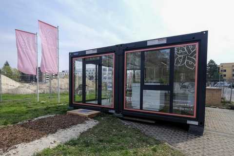 Containerbüro mit großen Fenstern, daneben drei rosa Fahnen im Freien.