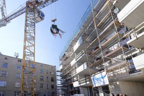 Kran hebt geschmückten Richtkranz neben Rohbau eines mehrstöckigen Gebäudes.