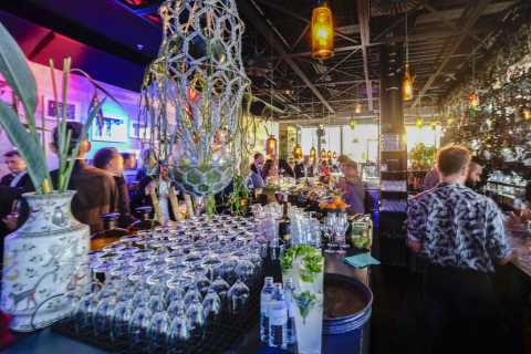 Bunte Bar mit vielen Gästen, Gläsern, Pflanzen und beleuchteter Dekoration.