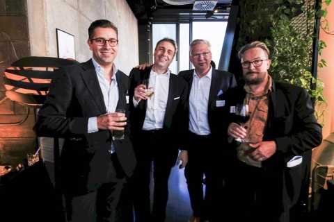 Vier Männer in Anzügen stehen zusammen und halten Getränke in der Hand.