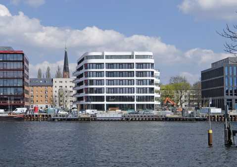 Modernes weißes Bürogebäude am Wasser, umgeben von weiteren Gebäuden.