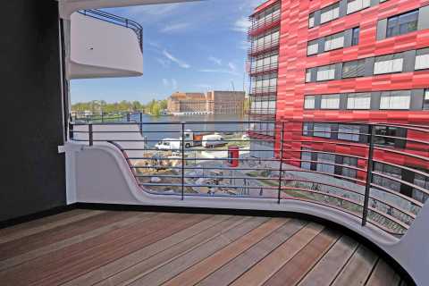 Moderner Balkon mit Holzboden, Blick auf Fluss und rot-graues Gebäude.
