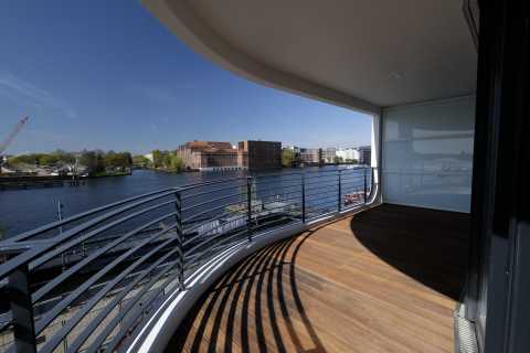 Großer, überdachter Balkon mit Blick auf einen Fluss und rote Backsteingebäude.