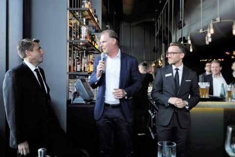 Drei Männer in Anzügen sprechen in einer Bar vor einem Publikum.