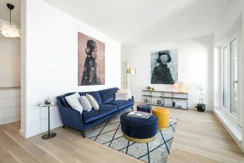 Modernes Wohnzimmer mit blauem Sofa, Porträts, Teppich und großen Fenstern.