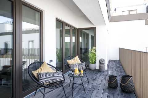 Moderne Balkonmöbel, Glastisch mit Getränken, Holzboden, helle Umgebung.