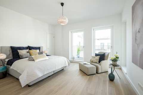 Helles Schlafzimmer mit Doppelbett, Sessel, großen Fenstern und moderner Deko.