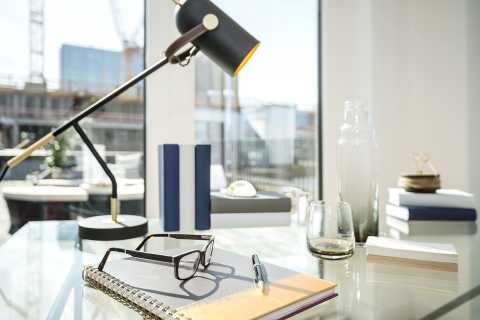 Heller Schreibtisch mit Lampe, Brille, Notizbuch, Stift und Glas vor Fenster.