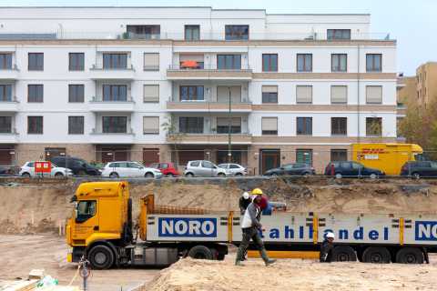 Bauarbeiter laufen vor einem gelben LKW an einer Baustelle vor Wohnhäusern.