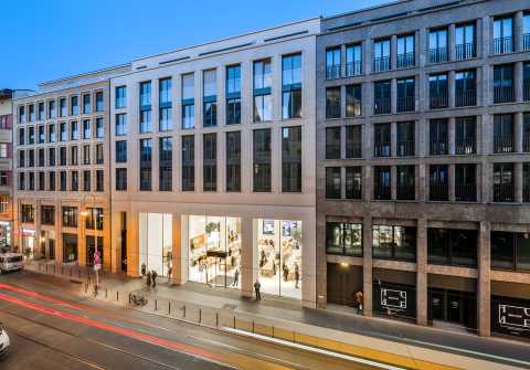 Belebte Straße mit modernen Gebäuden und beleuchtetem Schaufenster abends.
