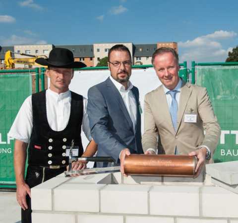 Drei Männer legen eine Kupferzeitkapsel bei einer Grundsteinlegung ein.